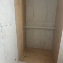 Wnętrze kabiny prysznicowej w trakcie prac wykończeniowych, widoczne płytki imitujące drewno na podłodze i ścianie, oraz jasne płytki ceramiczne na pozostałych ścianach i podłodze łazienki,...