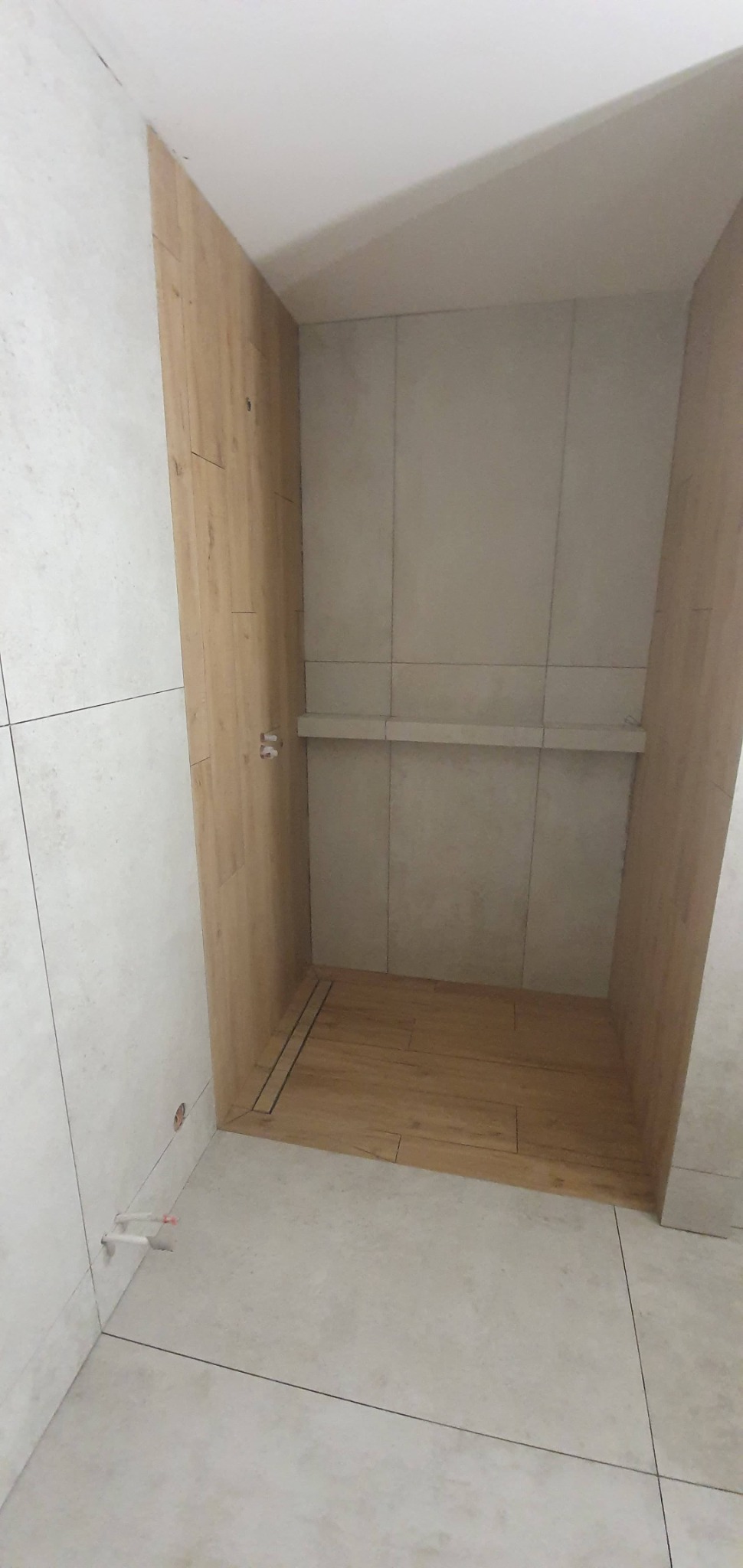 Wnętrze kabiny prysznicowej w trakcie prac wykończeniowych, widoczne płytki imitujące drewno na podłodze i ścianie, oraz jasne płytki ceramiczne na pozostałych ścianach i podłodze łazienki,...