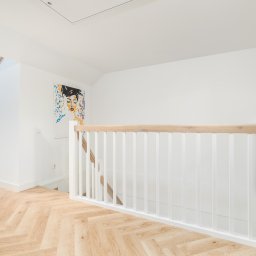 RSIFurniture - Jasne wnętrze z drewnianą podłogą w jodełkę, białymi ścianami i schodami z białą balustradą. Na ścianie obraz w stylu pop-art. Widoczne okno dachowe i roślina doniczkowa.