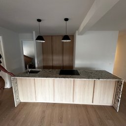 RSIFurniture - Jasna wyspa kuchenna z kamiennym blatem i płytą indukcyjną, oświetlona dwoma czarnymi lampami wiszącymi. W tle zabudowa kuchenna z drewnianymi frontami.