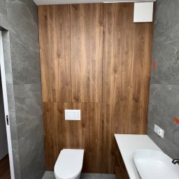 RSIFurniture - Nowoczesna łazienka z drewnianą zabudową szafek, szarą płytką imitującą kamień i białą armaturą. Minimalistyczny design, widoczny fragment umywalki i toalety.