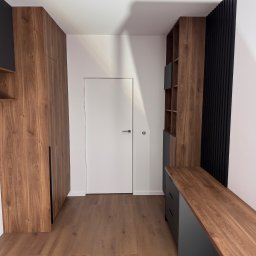 RSIFurniture - Nowoczesny przedpokój z drewnianą zabudową: szafa, regał z czarnymi akcentami i długi blat. Minimalistyczne drzwi i podłoga z paneli w ciepłym odcieniu drewna.