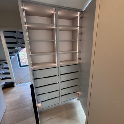 RSIFurniture - Szafa wnękowa z oświetleniem LED, półkami i szufladami, dopasowana do skosu pod schodami. Widoczna część wnętrza szafy i fragment schodów.