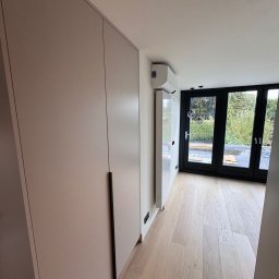 RSIFurniture - Jasna szafa na wymiar z długimi, czarnymi uchwytami w nowoczesnym wnętrzu z drewnianą podłogą i dużymi oknami balkonowymi z czarnymi ramami.