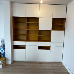 RSIFurniture - Biało-żółta szafa wnękowa z półkami i szafkami, o nieregularnym układzie, w jasnym pomieszczeniu z drewnianą podłogą. Widoczna żarówka na suficie.