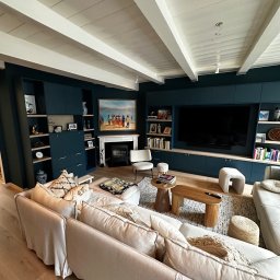 RSIFurniture - Elegancki salon z zabudową meblową w odcieniach granatu, beżową sofą i drewnianymi akcentami. Na ścianie obraz nad kominkiem. Białe belki stropowe.