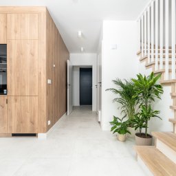RSIFurniture - Jasne wnętrze domu z drewnianą zabudową kuchenną, schodami i roślinami doniczkowymi. Minimalistyczny design, białe ściany i podłoga. Widok na korytarz z ciemnymi drzwiami.