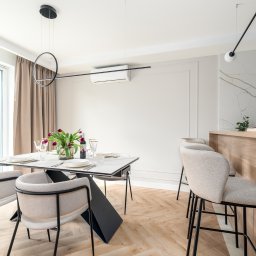 RSIFurniture - Jasna jadalnia z parkietem w jodełkę. Stół z nakryciem, krzesła z beżową tapicerką. W tle okno z widokiem na zieleń i marmurowa ściana z drewnianym blatem.