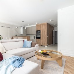 RSIFurniture - Jasne, przestronne wnętrze: salon z beżową sofą, okrągłym stolikiem kawowym i widokiem na kuchnię z drewnianą zabudową. Parkiet w jodełkę, nowoczesne oświetlenie.