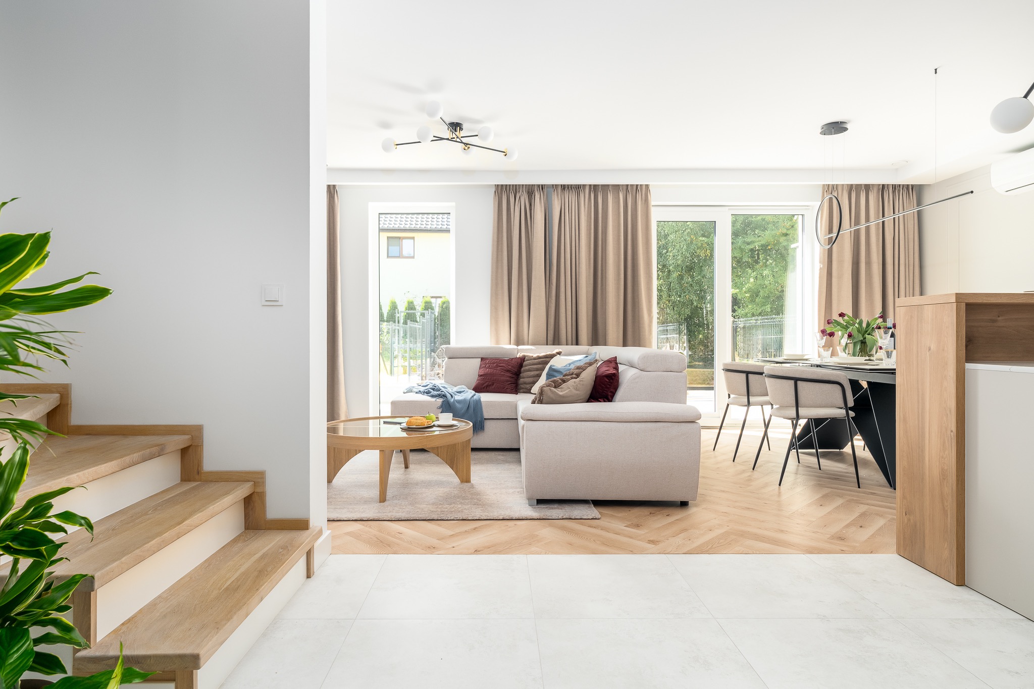 Jasne, przestronne wnętrze domu z drewnianymi schodami, beżową sofą i stołem jadalnym. Parkiet w jodełkę, duże okna z widokiem na zieleń. Nowoczesne oświetlenie.