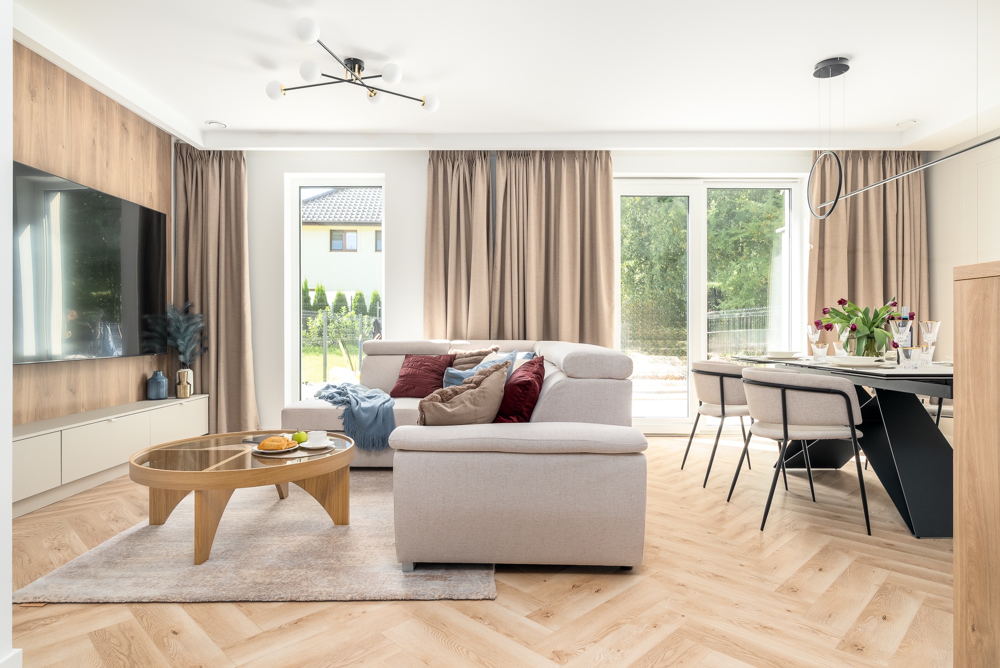 Jasne wnętrze salonu z drewnianą podłogą w jodełkę, beżową sofą z poduszkami, stolikiem kawowym i stołem z krzesłami. Widok na ogród przez okna.