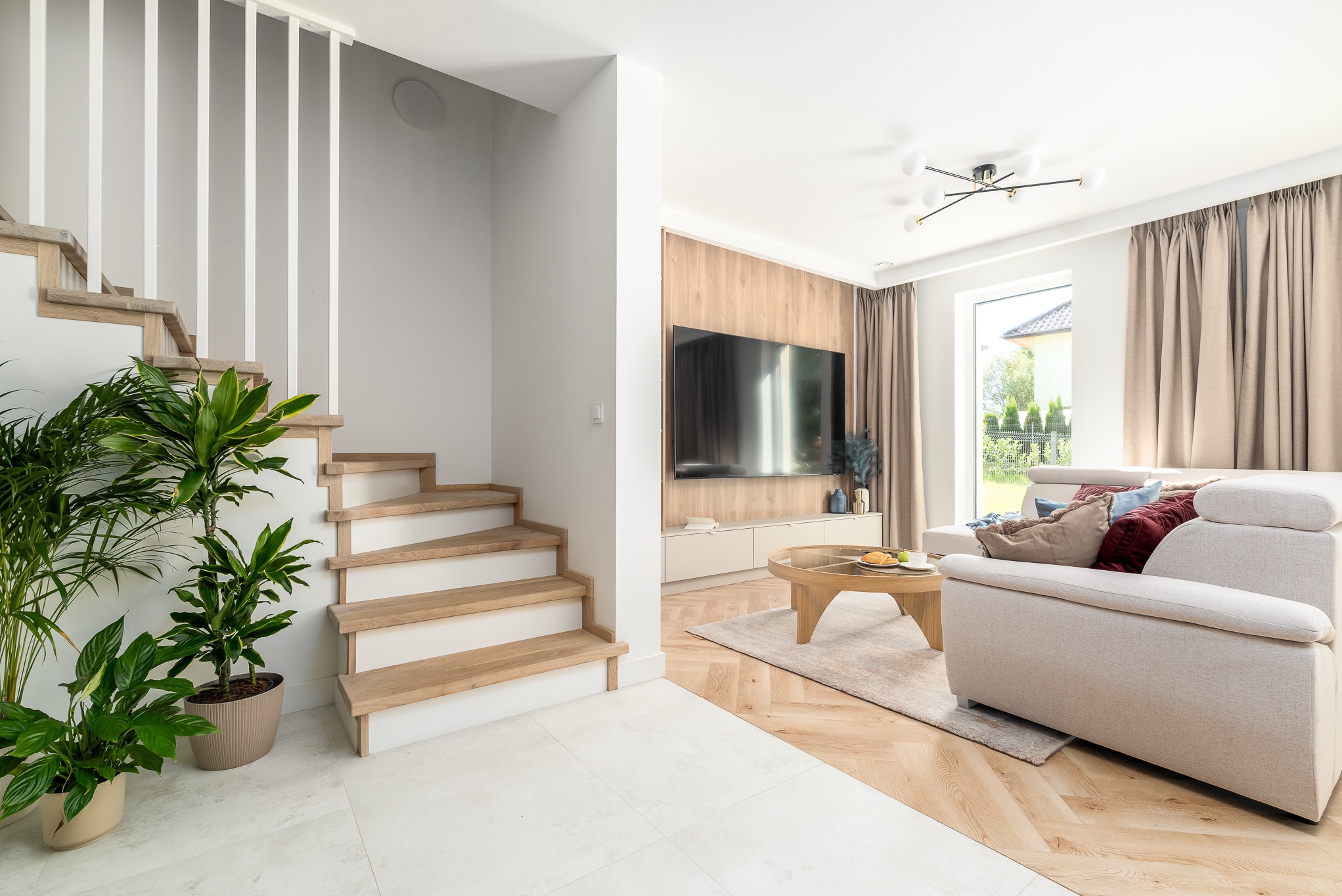 Jasne wnętrze domu: drewniane schody z roślinami, salon z telewizorem i beżową sofą. Minimalistyczny design, naturalne światło, nowoczesne oświetlenie.