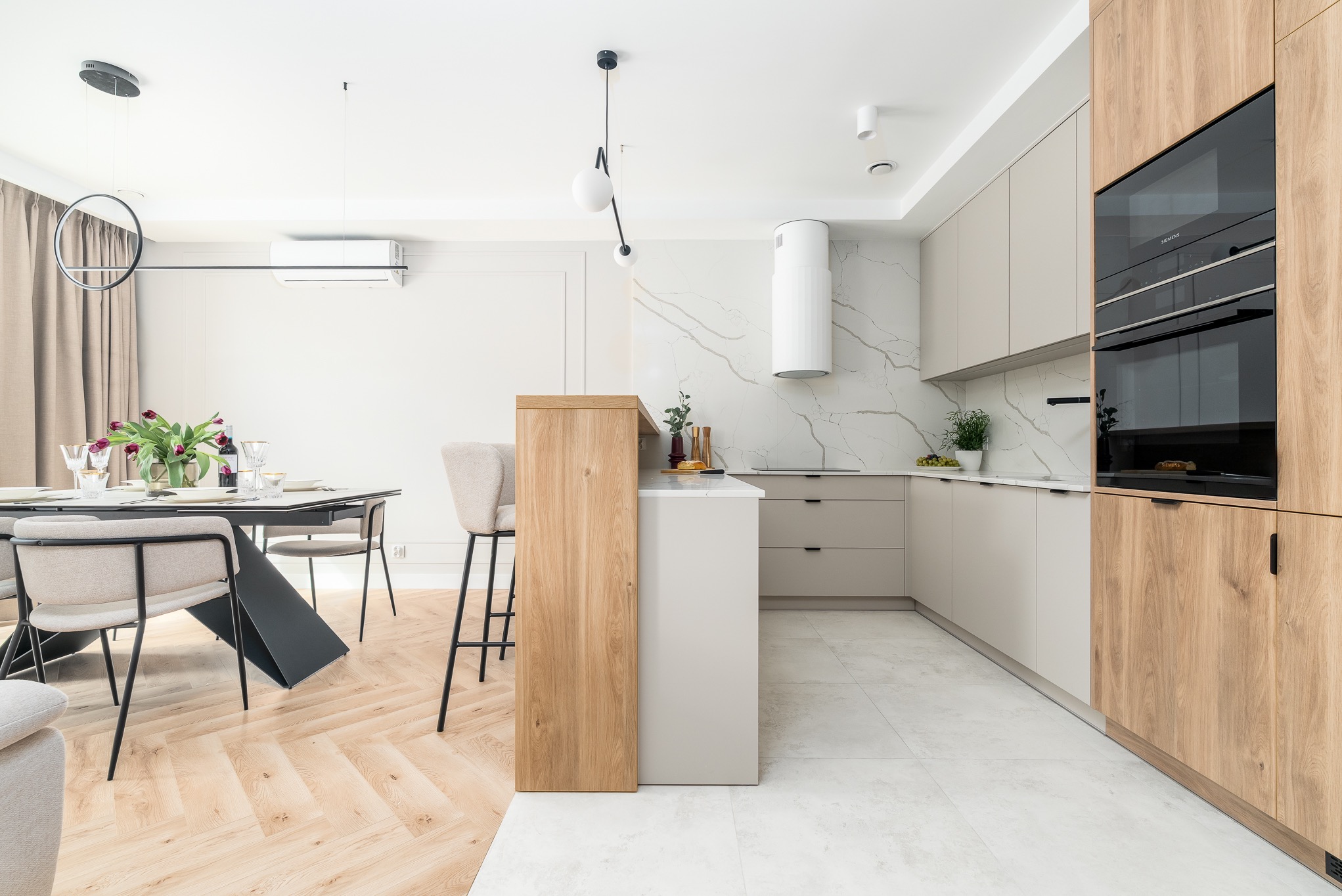 Jasna kuchnia z drewnianymi akcentami: wyspa, szafki, sprzęt AGD. Marmurowa ściana, parkiet w jodełkę, stół z kwiatami. Nowoczesny design, minimalistyczny styl.