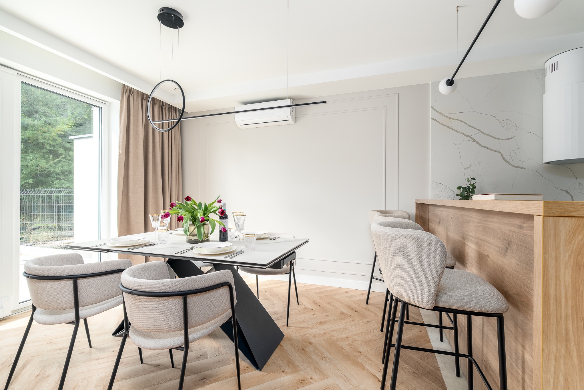 Jasna jadalnia z parkietem w jodełkę: stół z nakryciem, krzesła z beżową tapicerką, kwiaty w wazonie. W tle okno i marmurowa ściana z drewnianym blatem.