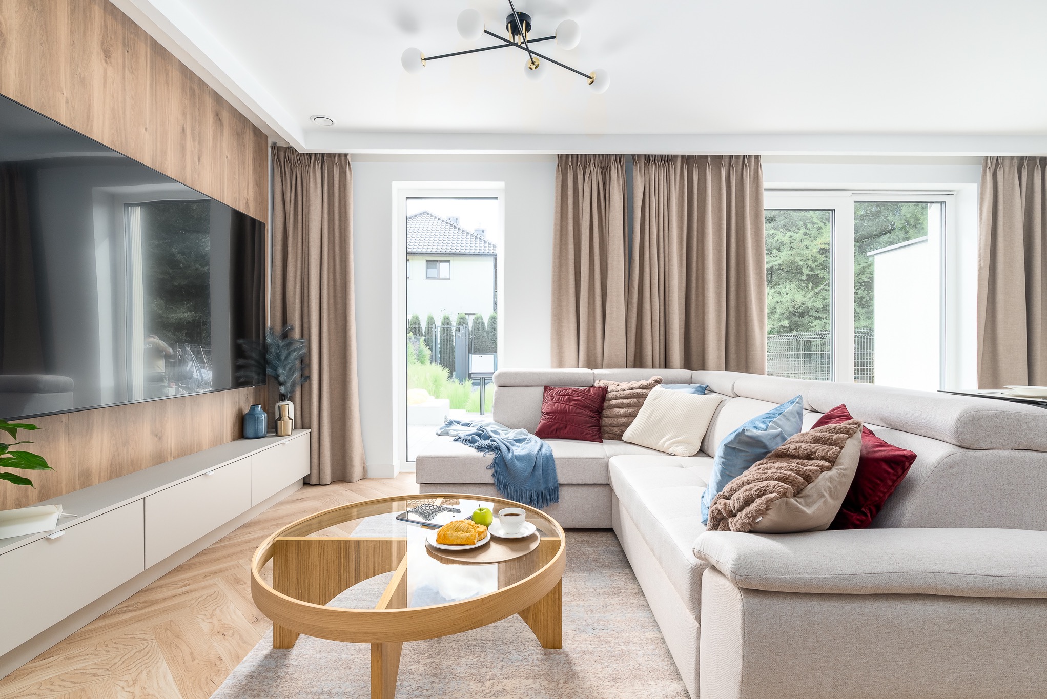 Jasny salon z narożnikiem, okrągłym stolikiem i drewnianą ścianą TV. Widok na ogród przez okno. Nowoczesne oświetlenie i beżowe zasłony.