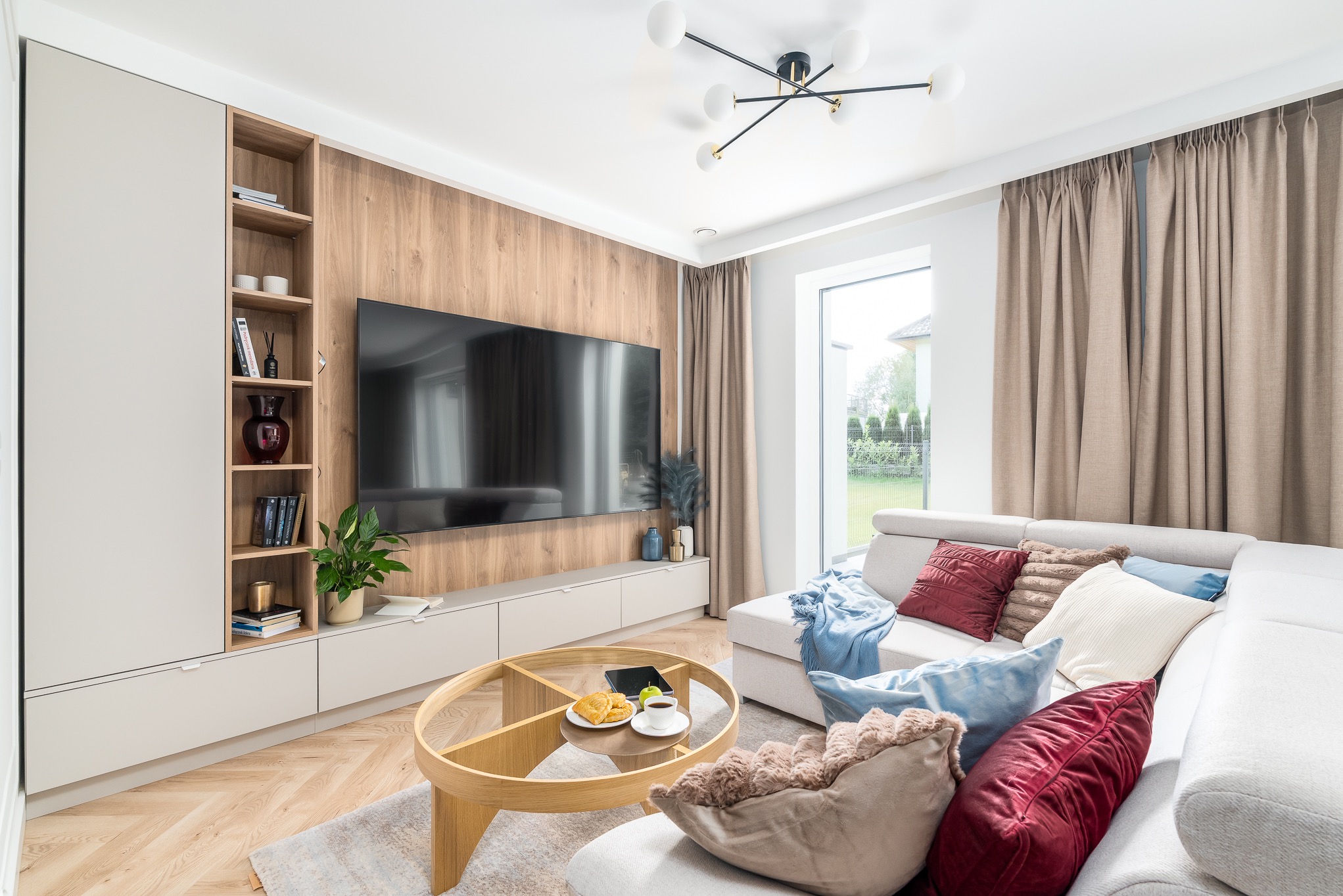 Nowoczesny salon z zabudową meblową: szafa z półkami, szafka RTV pod telewizorem. Okrągły stolik kawowy z przekąskami. Jasna sofa z poduszkami i pledem.