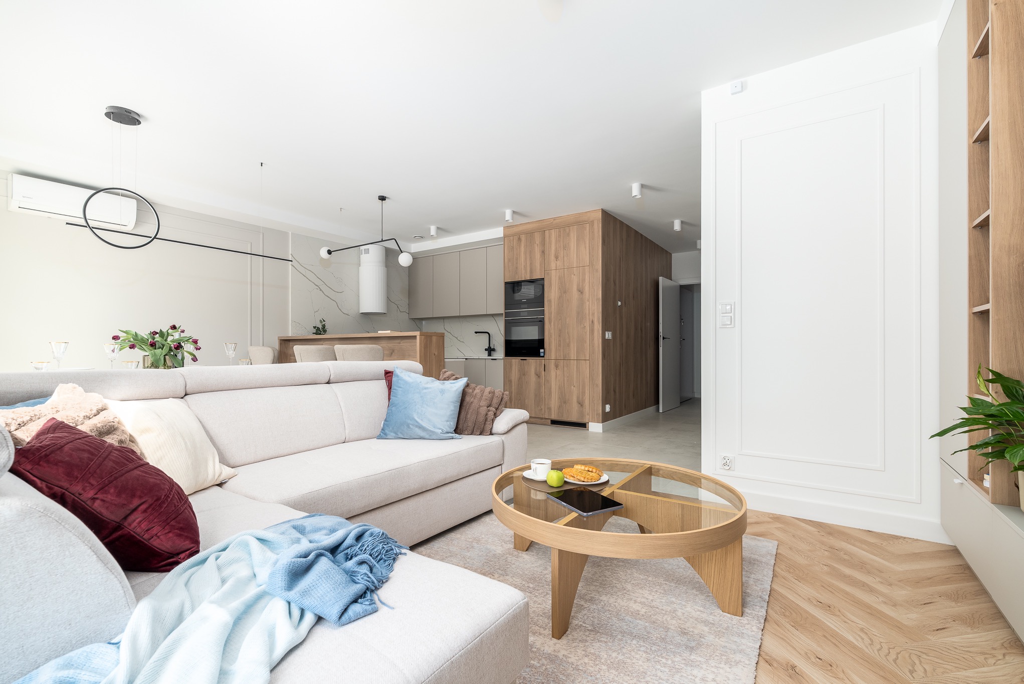 Jasne wnętrze: sofa z poduszkami, okrągły stolik z przekąskami, kuchnia z drewnianymi frontami w tle. Minimalistyczny design, parkiet w jodełkę.