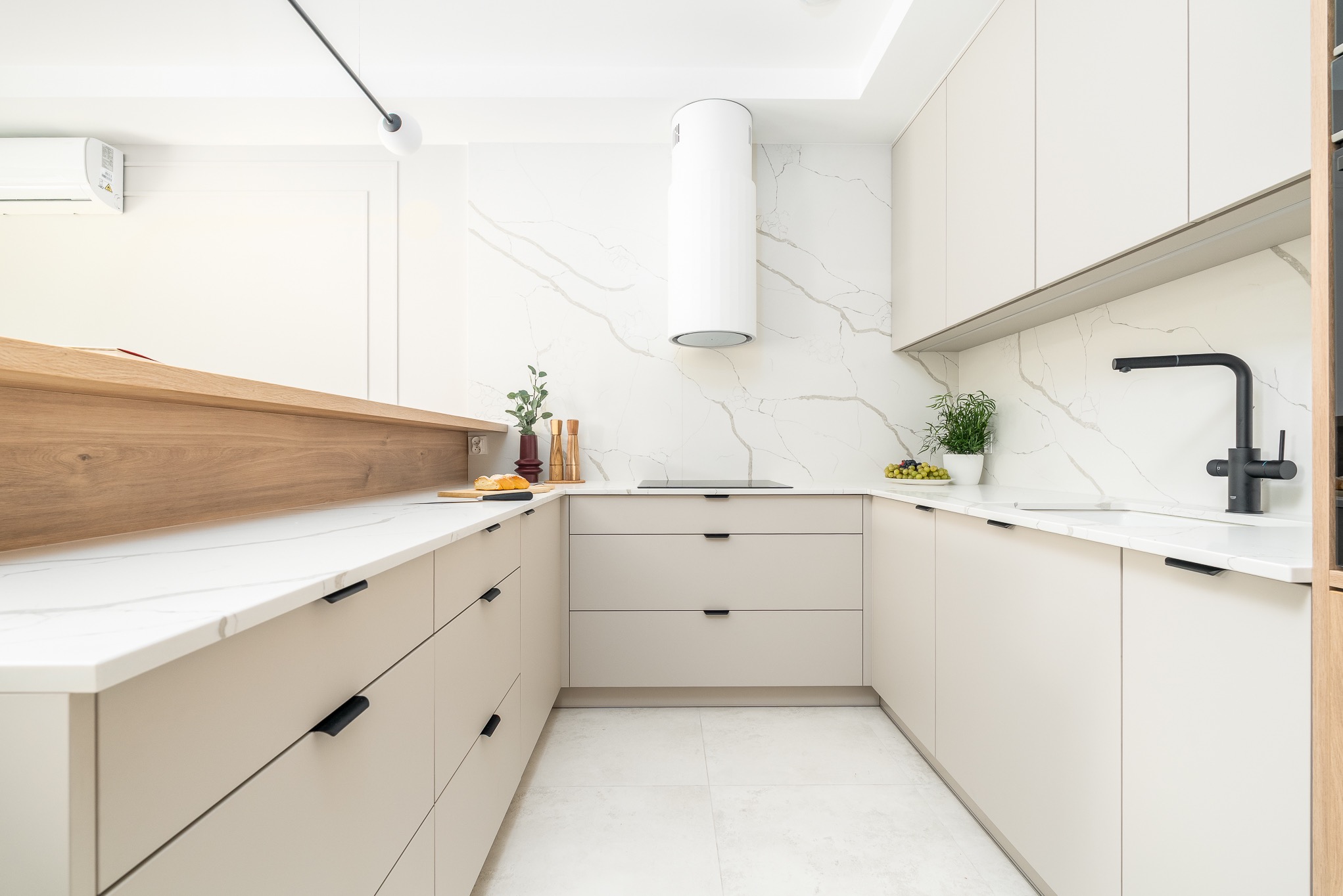Jasna kuchnia w kształcie litery U z beżowymi szafkami, czarnymi uchwytami i blatem imitującym marmur. Na blacie owoce i zioła w doniczce. Minimalistyczny design.