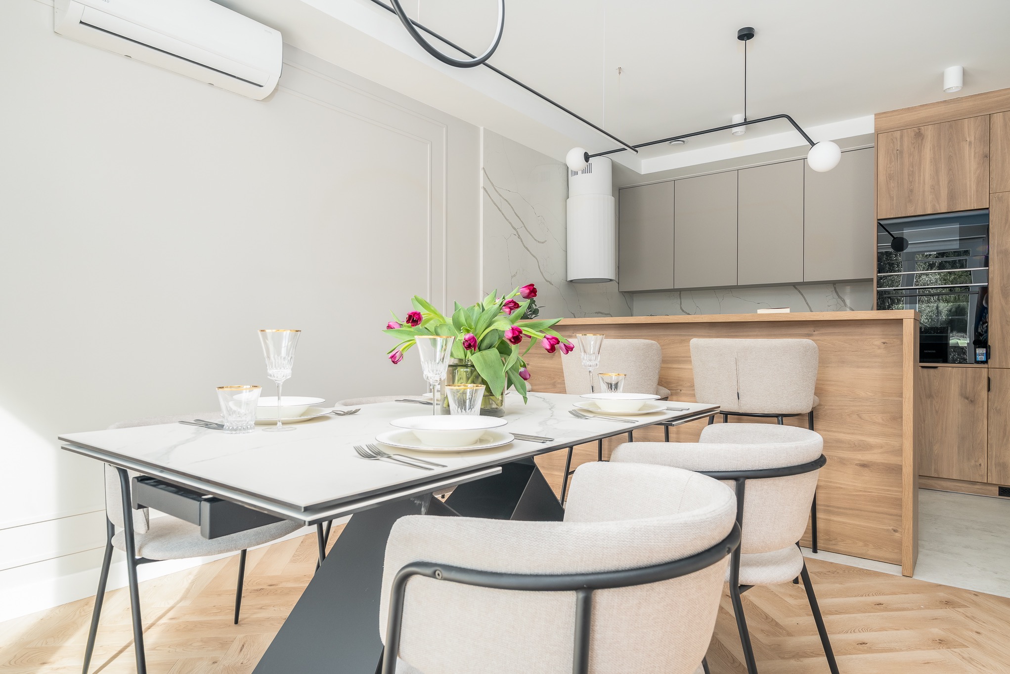 Jasna jadalnia z marmurowym stołem, kwiatami i beżowymi krzesłami. W tle zabudowa kuchenna z drewnianymi frontami i sprzęt AGD. Nowoczesne oświetlenie.