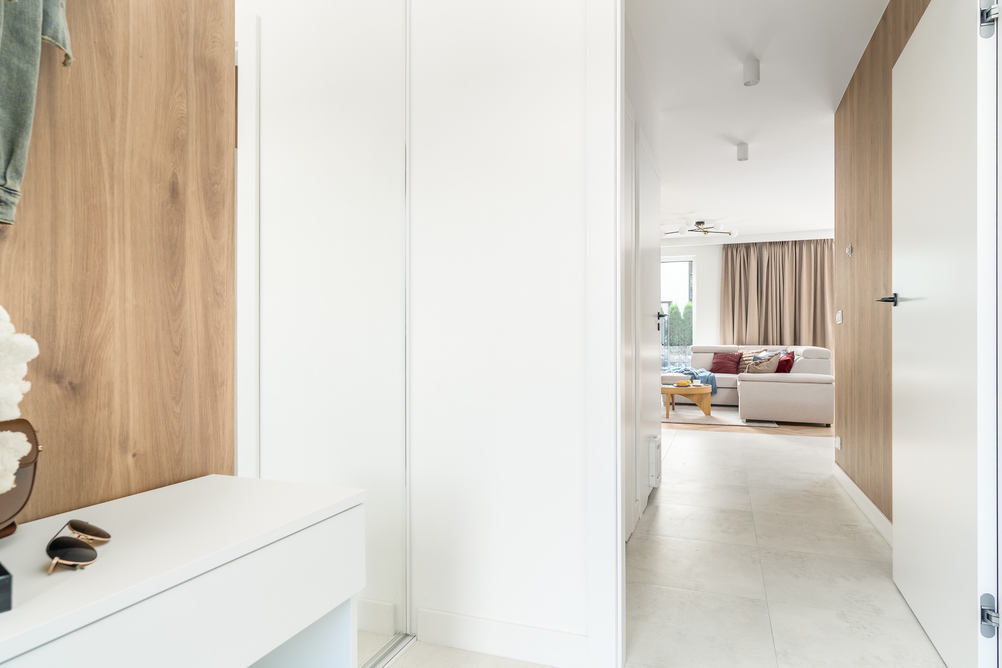 Biała szafa wnękowa z lustrzanymi drzwiami w przedpokoju, uzupełniona drewnianą boazerią i komodą z okularami. W tle widoczny salon z sofą.