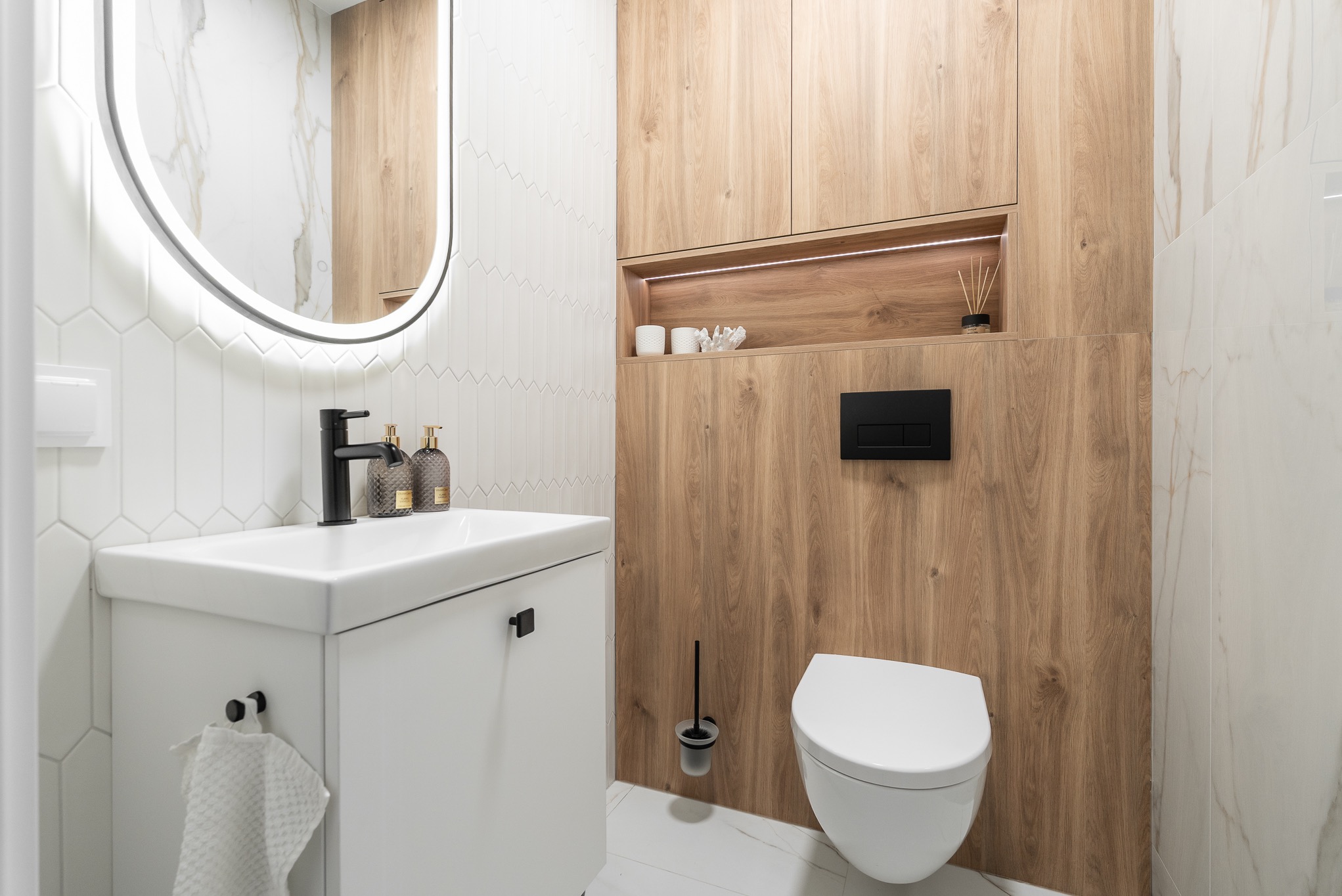 Nowoczesna łazienka z drewnianą zabudową, owalnym lustrem z podświetleniem LED, białą umywalką i toaletą. Minimalistyczny design, jasne płytki i czarne akcenty.