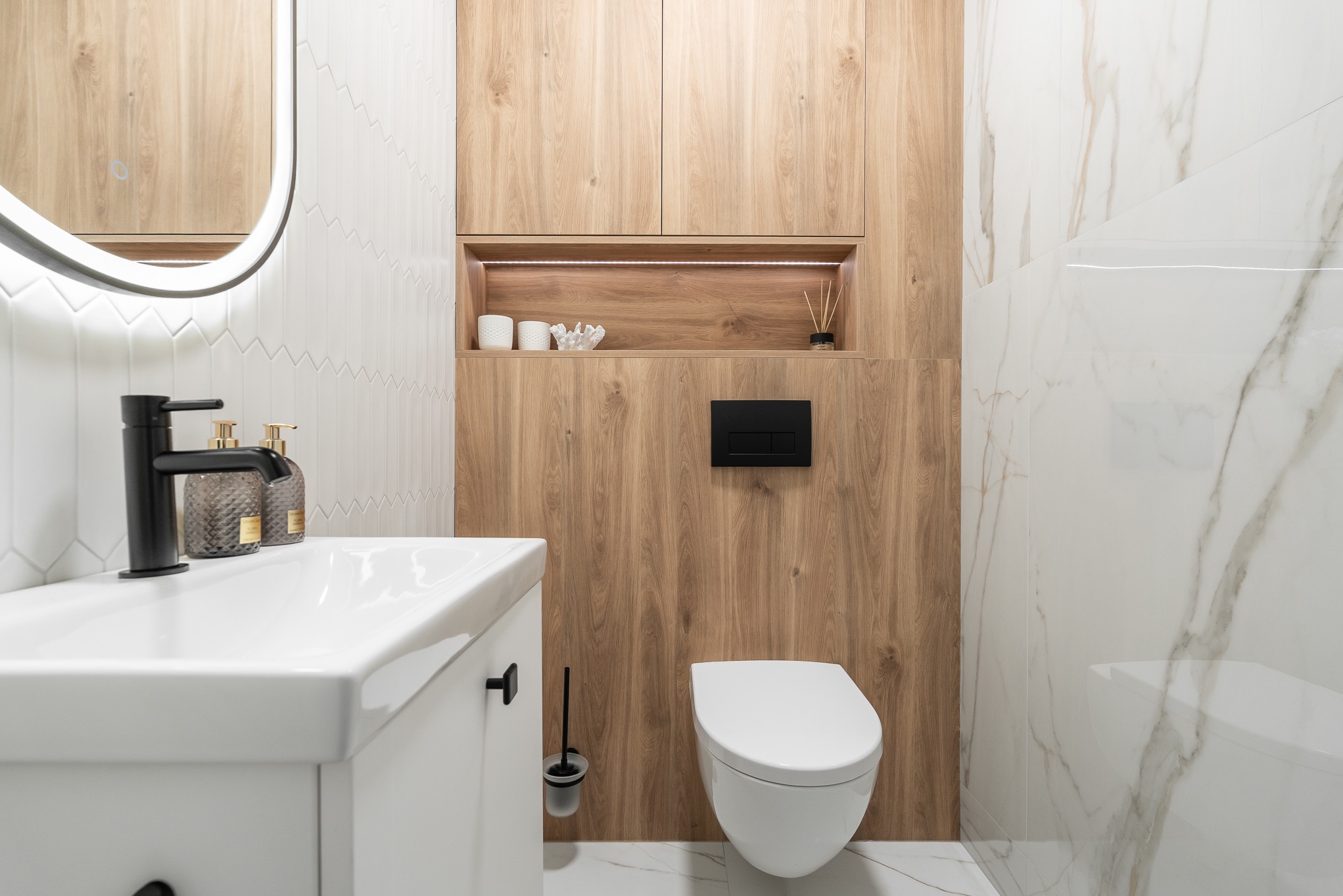 Nowoczesna łazienka z drewnianą zabudową, białą armaturą i geometrycznymi płytkami. Umywalka z czarną baterią, podświetlana wnęka z dekoracjami.