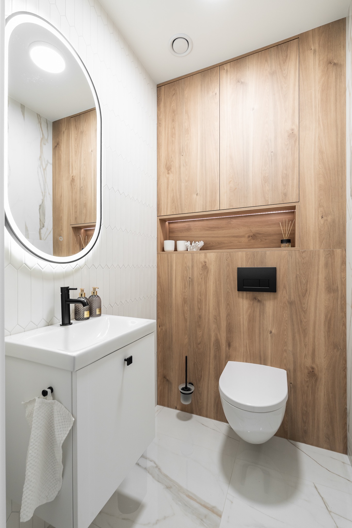 Nowoczesna łazienka z drewnianą zabudową, owalnym lustrem z podświetleniem LED, białą umywalką i toaletą. Minimalistyczny design, płytki imitujące marmur.