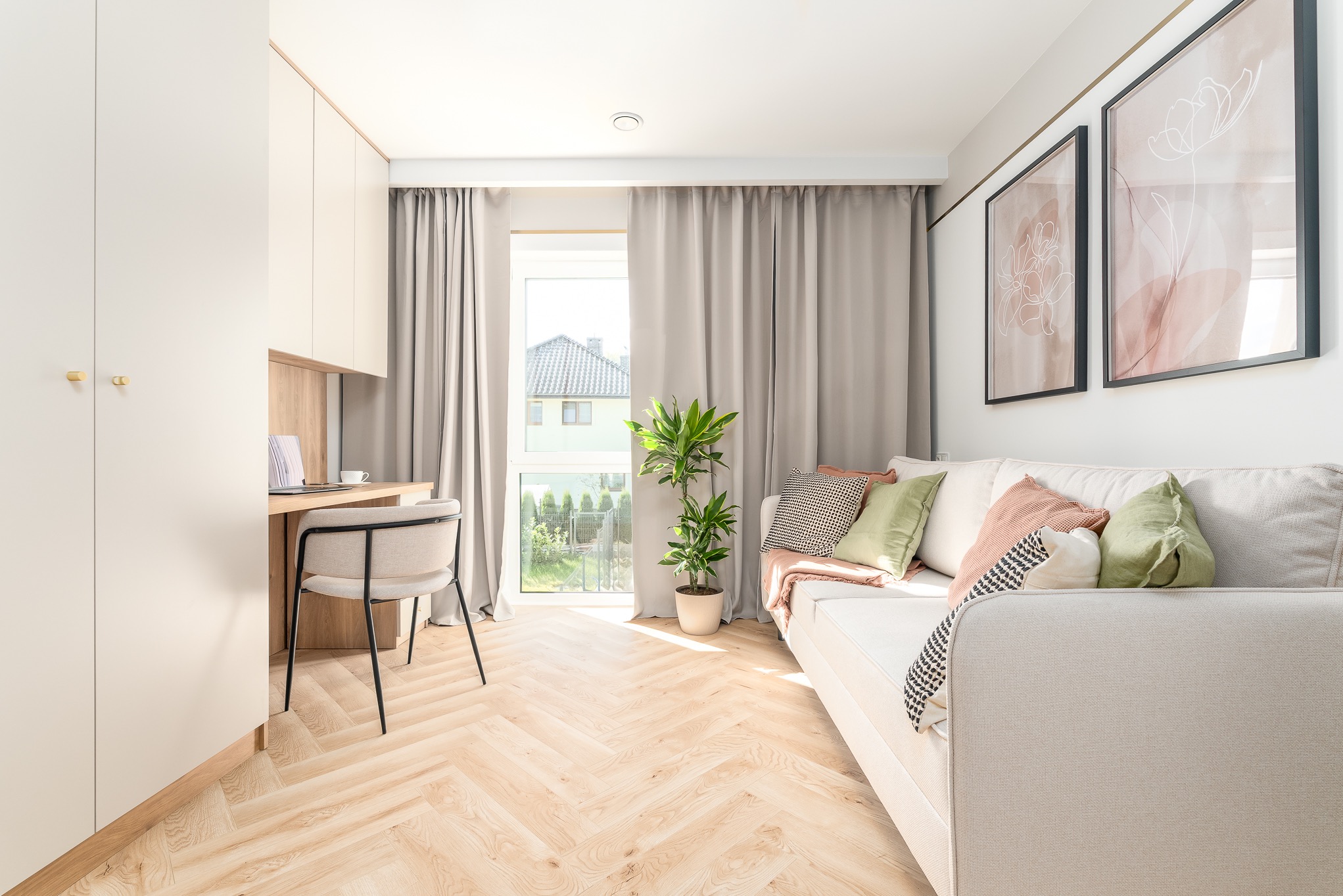 Jasne wnętrze z biurkiem, szafą i sofą. Parkiet w jodełkę, roślina w doniczce przy oknie. Dwa obrazy na ścianie nad sofą. Minimalistyczny design.