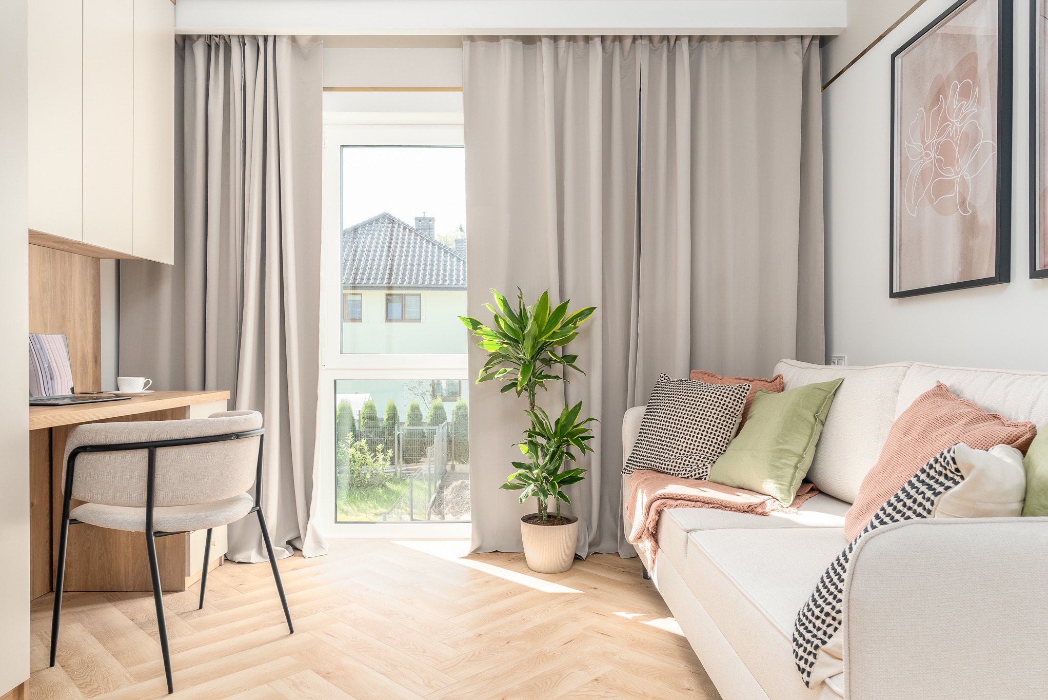Jasne wnętrze z biurkiem, sofą i oknem z widokiem na ogród. Neutralna kolorystyka, drewniana podłoga w jodełkę, roślina doniczkowa. Minimalistyczny design.
