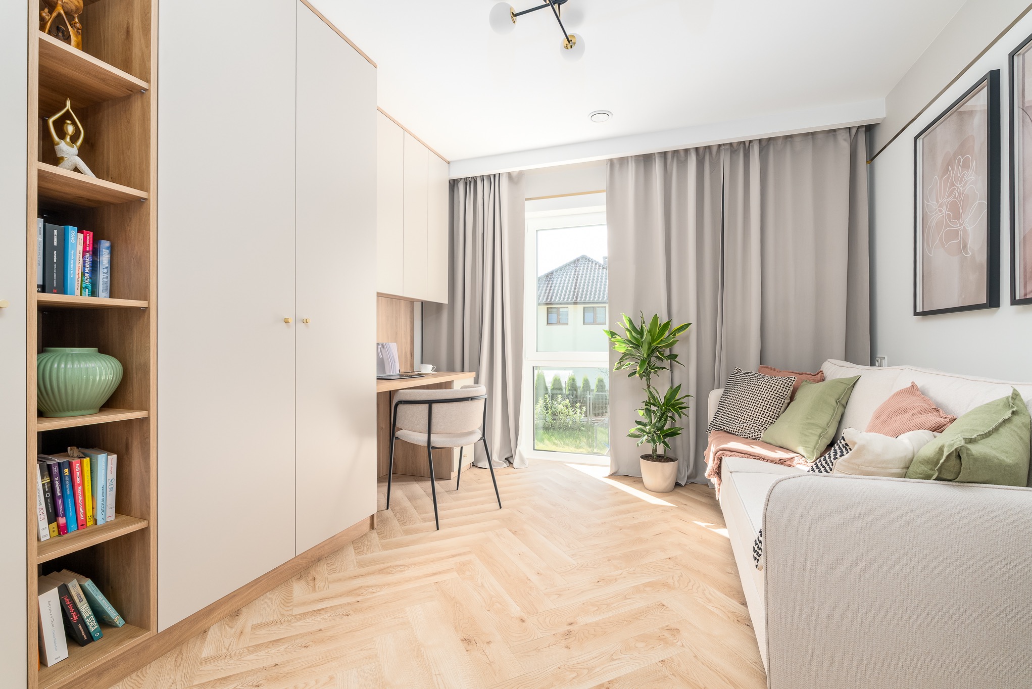 Jasne wnętrze pokoju z biurkiem, szafą w zabudowie, regałem z książkami i sofą. Parkiet w jodełkę, okno z zasłonami. Minimalistyczny design z akcentami roślinnymi.