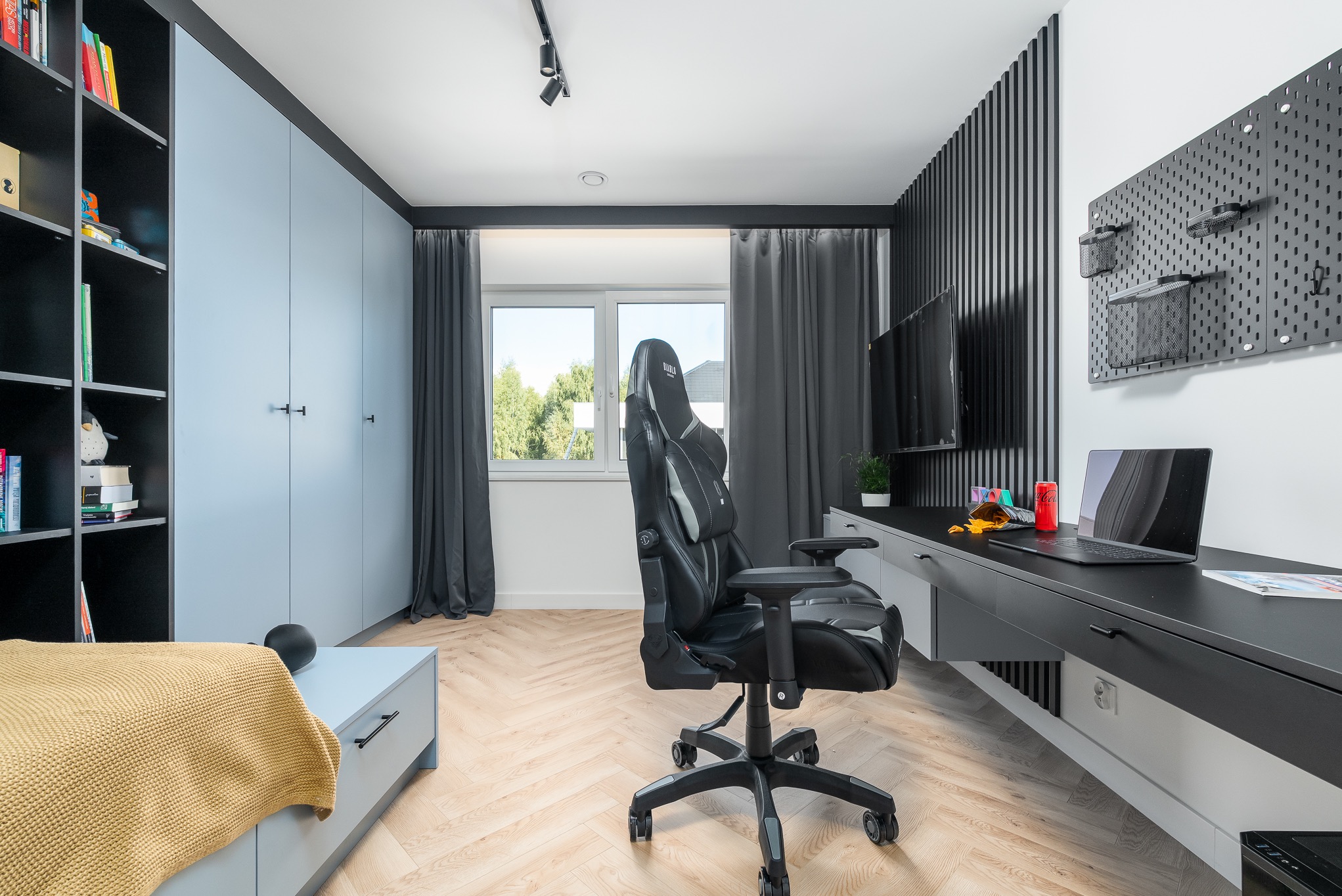 Pokój młodzieżowy z biurkiem, szafą i regałem w odcieniach szarości i błękitu. Na biurku laptop i napój. Krzesło gamingowe. Parkiet w jodełkę. Minimalistyczny design.