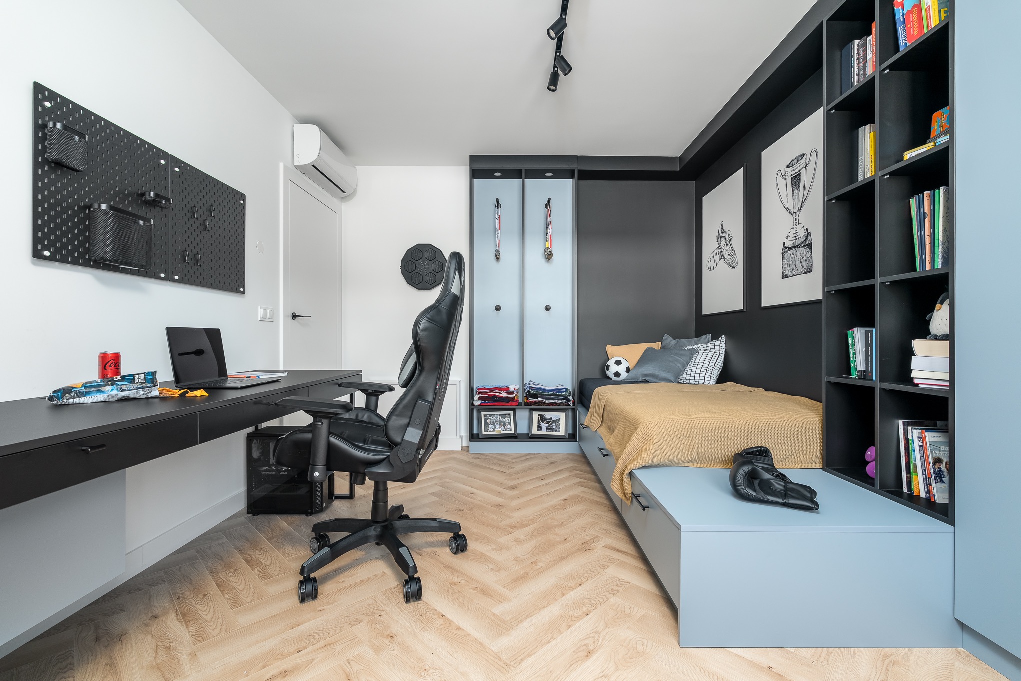 Pokój młodzieżowy z biurkiem, krzesłem gamingowym i łóżkiem z szafkami. Na ścianie tablica narzędziowa i grafiki sportowe. Parkiet w jodełkę. Nowoczesny design.