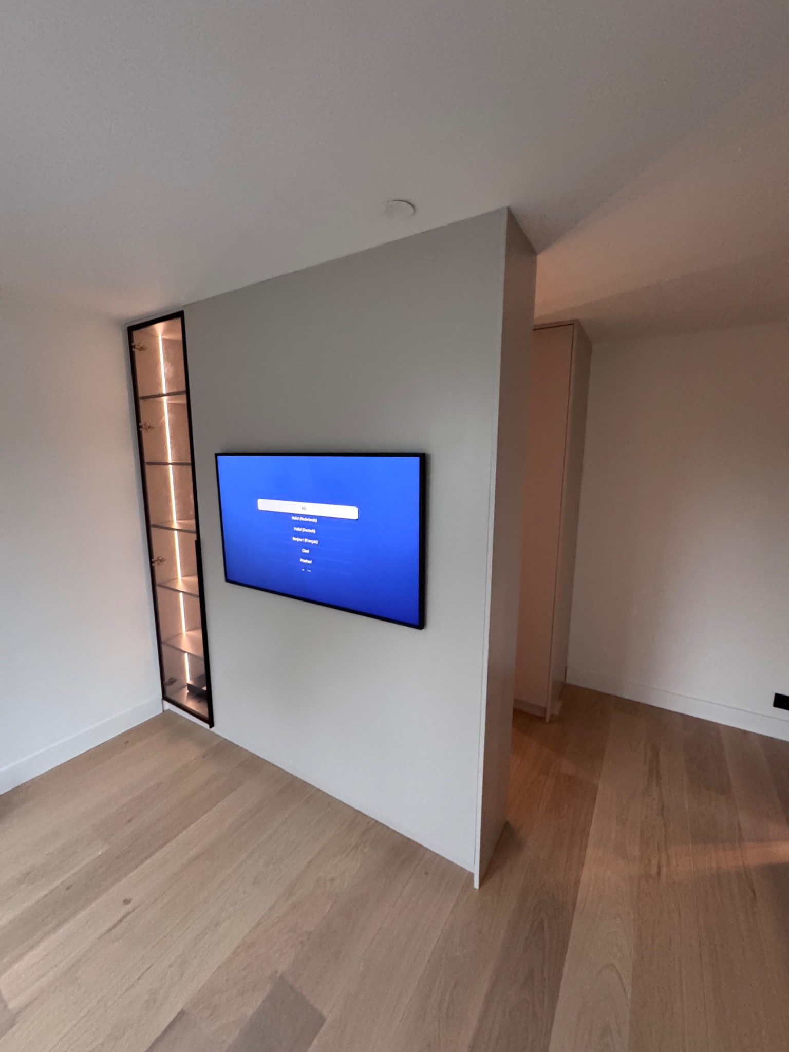 Nowoczesny salon z drewnianą podłogą, wbudowaną, podświetlaną szafką i telewizorem na ścianie. Minimalistyczny design, jasne kolory, eleganckie wykończenie.