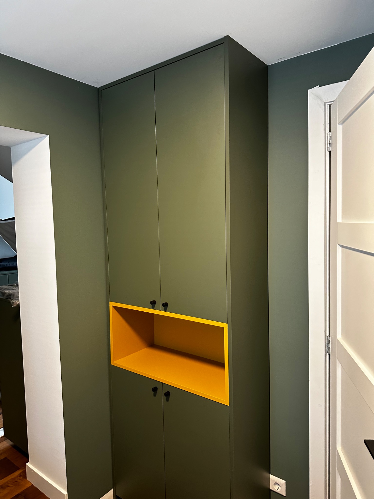 Wysoka szafa wnękowa z żółtą wnęką w Szczecinie, dopasowana kolorystycznie do ścian. Minimalistyczne czarne uchwyty, białe drzwi po prawej stronie.