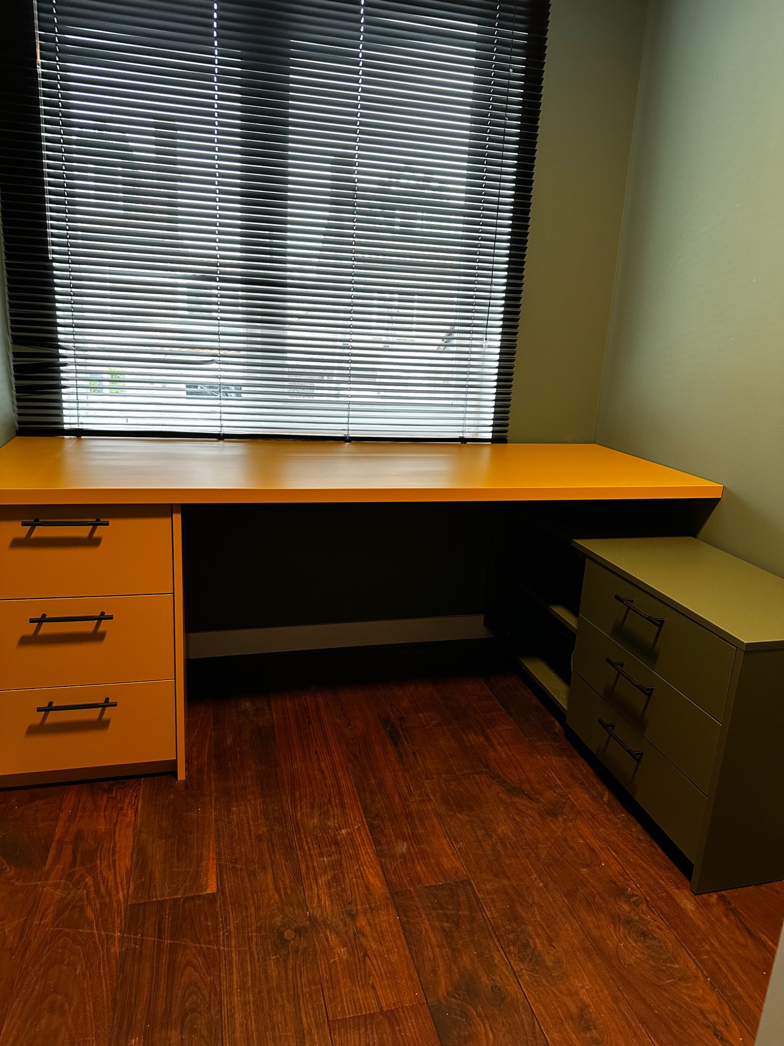 Nowoczesne biurko z szufladami w odcieniach pomarańczy i zieleni, ustawione przy oknie z żaluzjami. Parkiet z ciemnego drewna. Minimalistyczny design.