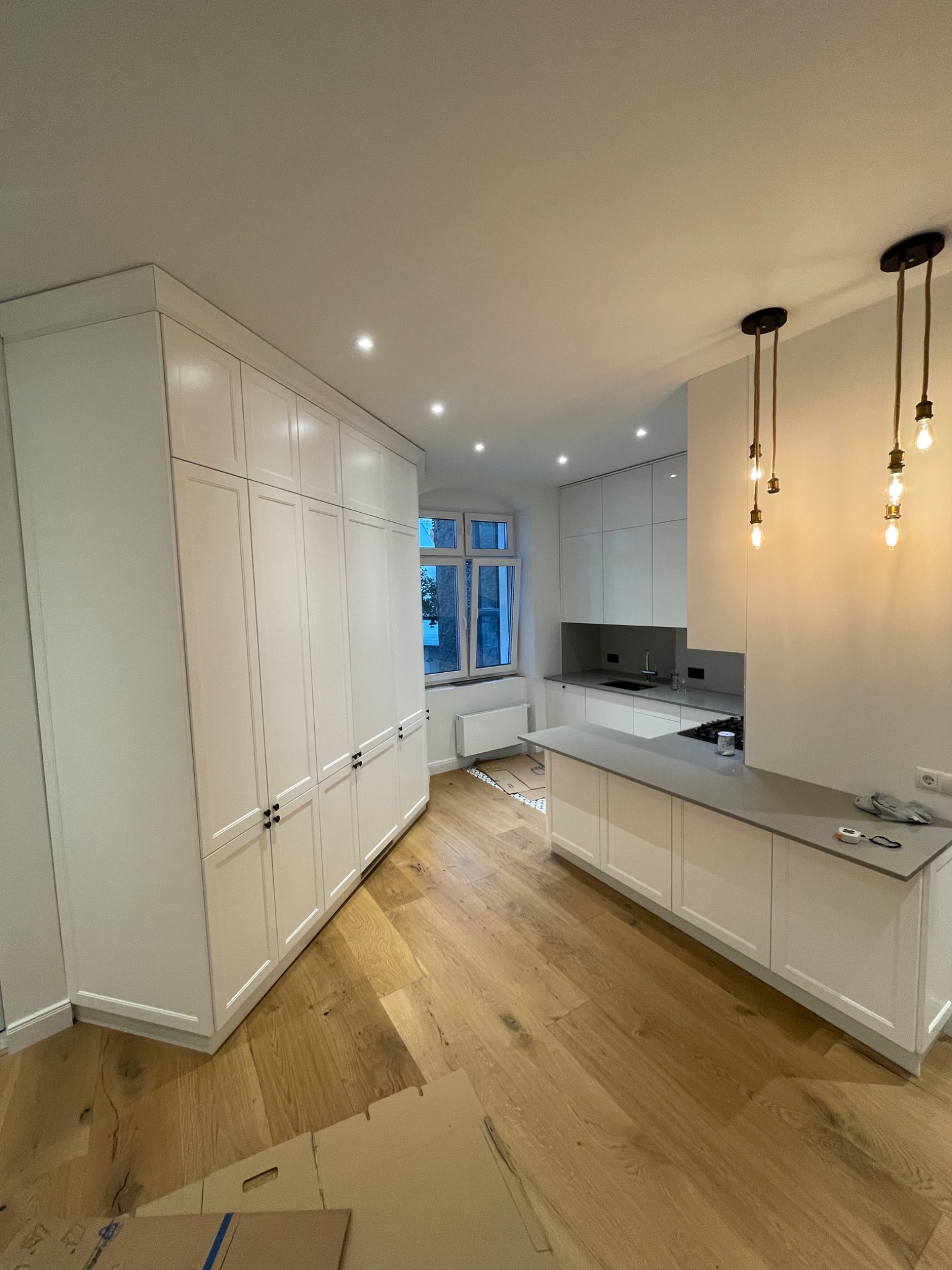 Jasna kuchnia z wyspą i szafą w zabudowie, drewniana podłoga. Minimalistyczny design z wiszącymi lampami. Widok na okno zza blatu.