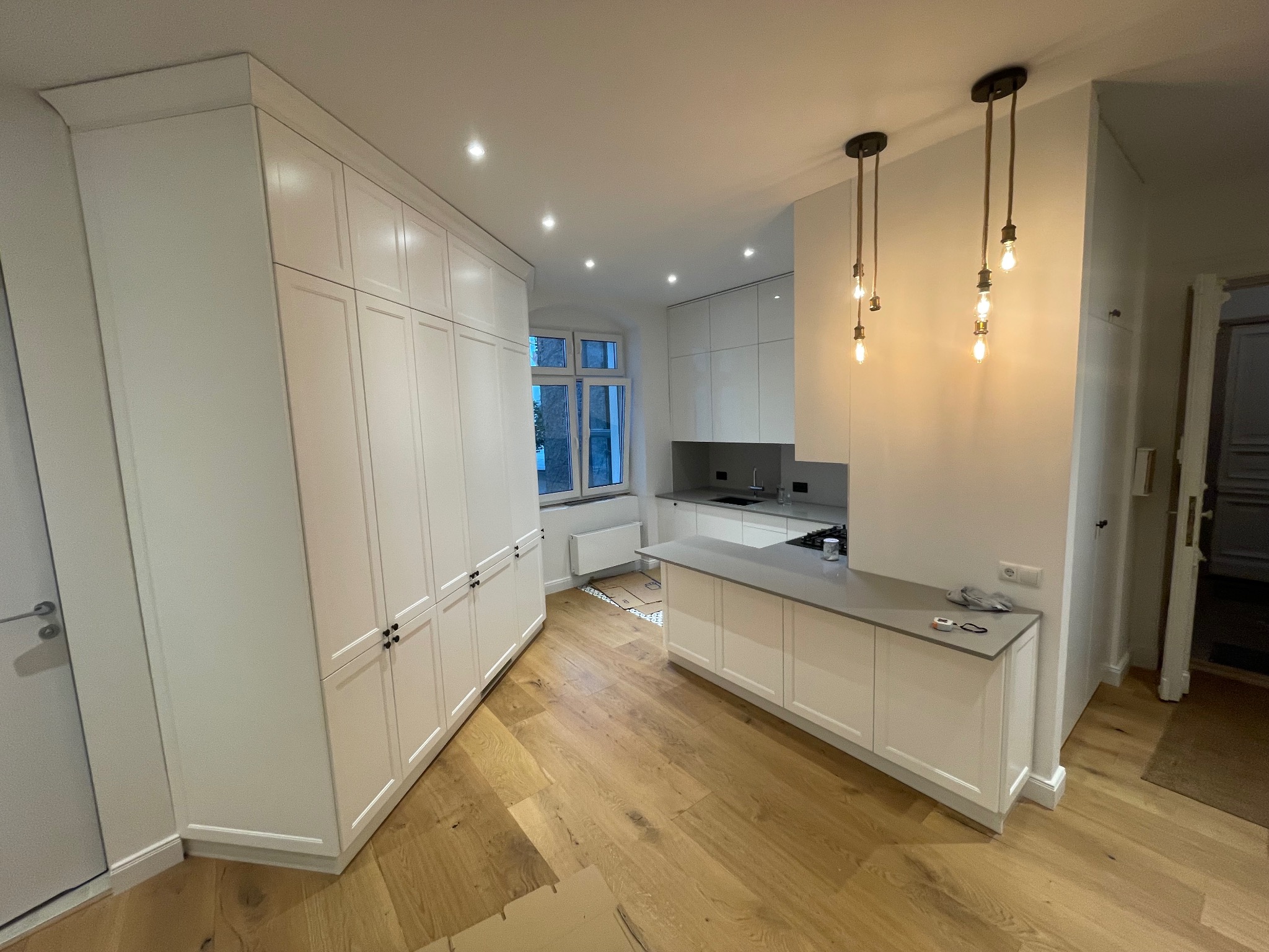 Jasna kuchnia z białymi szafkami na wymiar, drewnianą podłogą i szarym blatem. Widoczne wiszące lampy i okno. Minimalistyczny design.