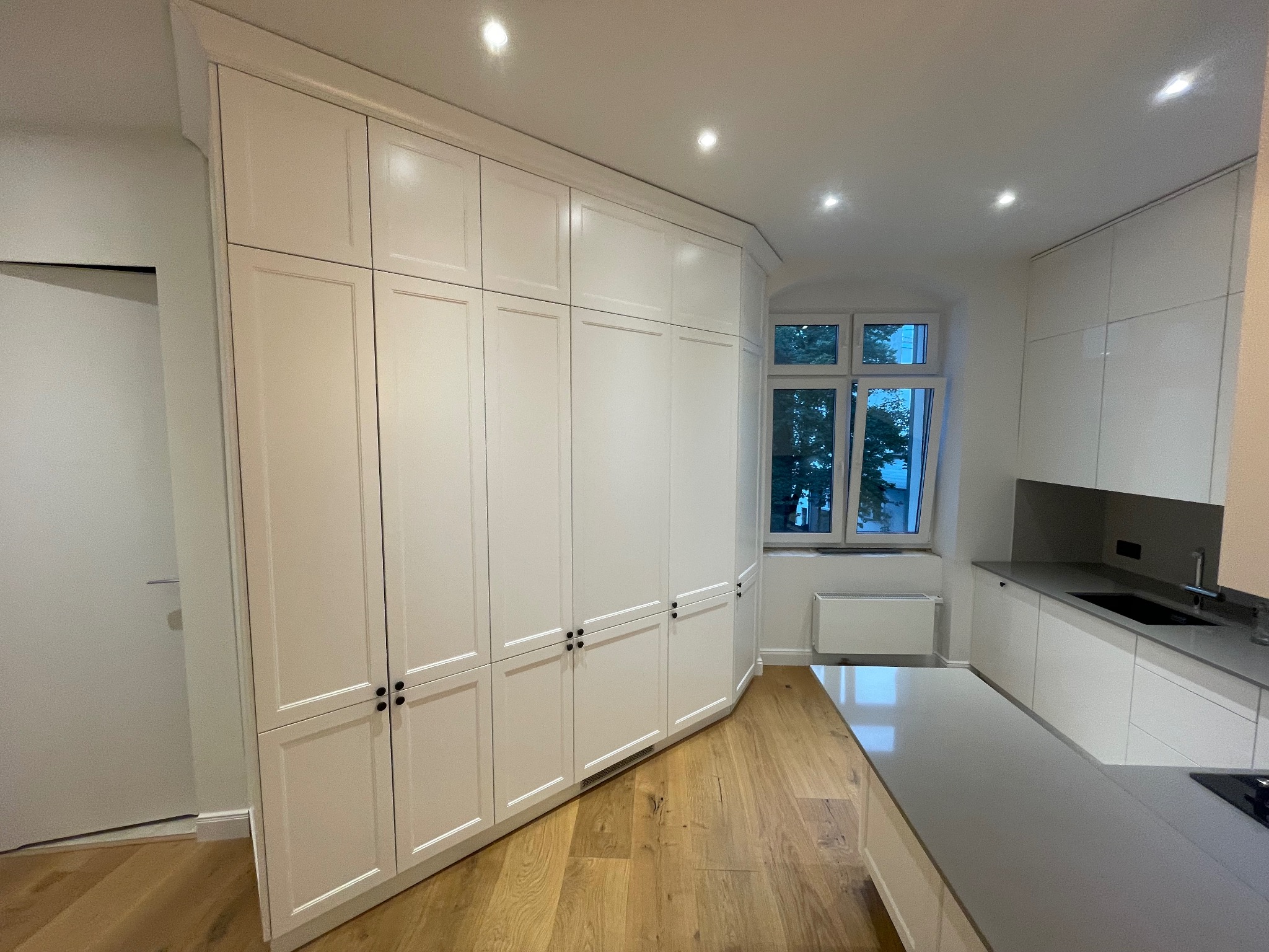 Biała zabudowa kuchenna z szafkami pod sufit, drewniana podłoga, szary blat i okno z widokiem na zieleń. Nowoczesne wykończenie kuchni.