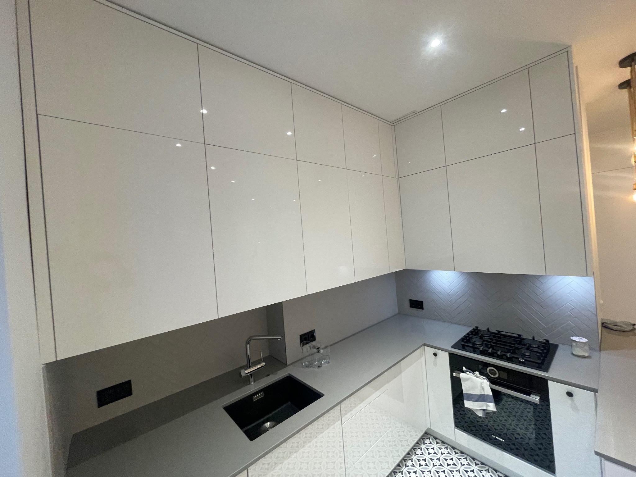 Biała, nowoczesna kuchnia z błyszczącymi frontami szafek, szarym blatem i wzorzystą podłogą. Uwagę przykuwa minimalistyczny zlew i płyta grzewcza.