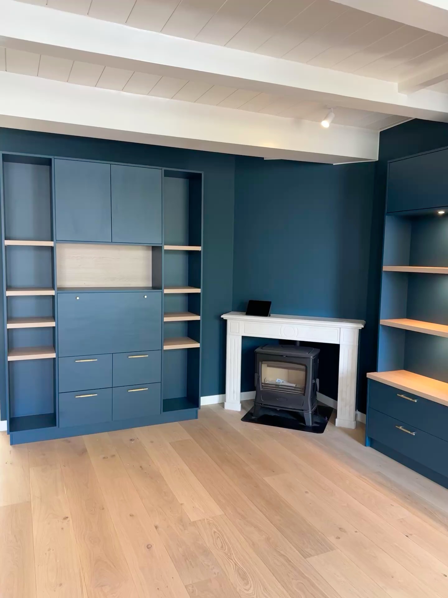 Niebieska zabudowa meblowa z jasnymi półkami i złotymi uchwytami, kominek z czarnym piecem, drewniana podłoga, biały sufit z belkami. Nowoczesne wnętrze.