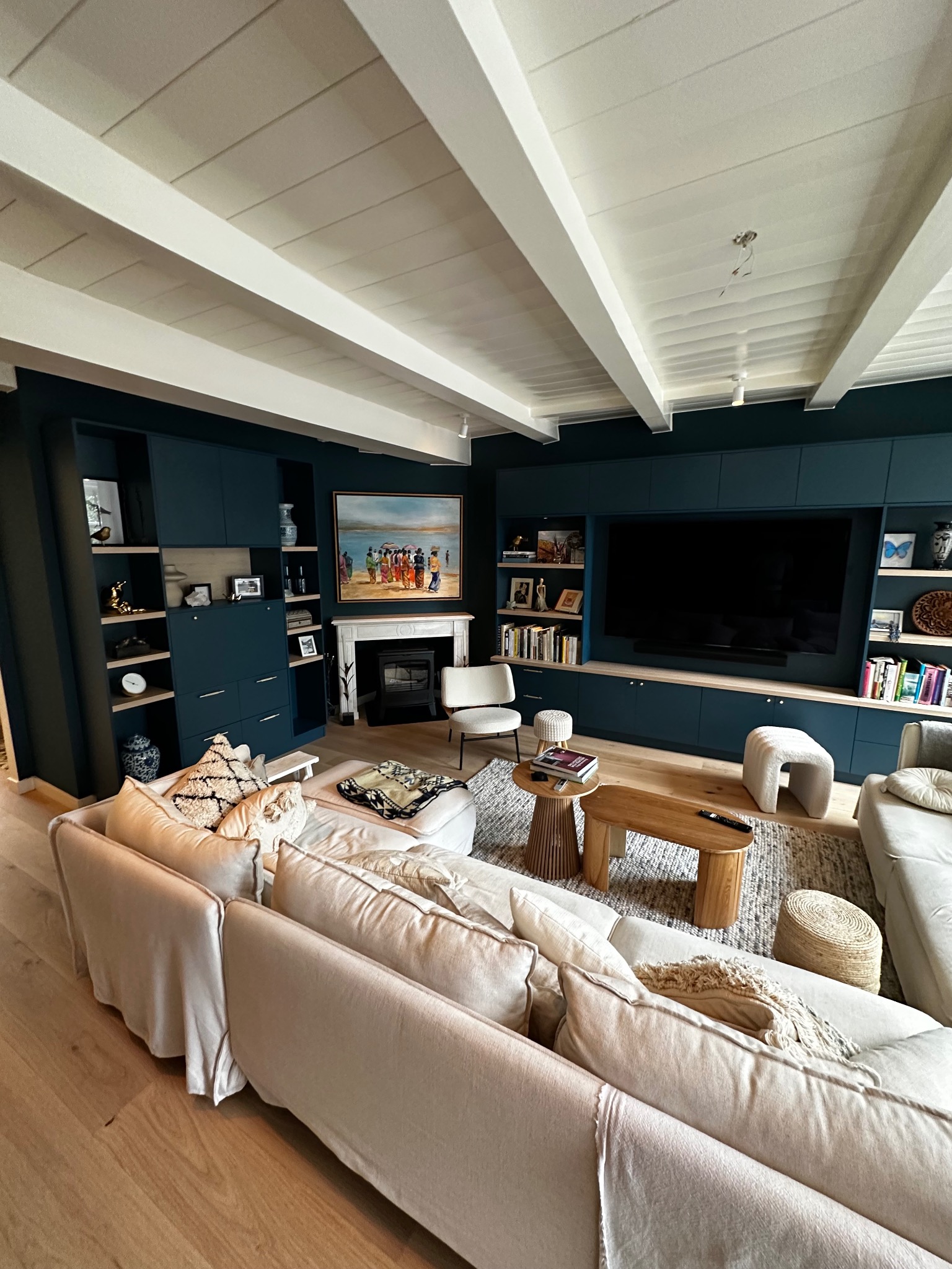 Elegancki salon z zabudową meblową w odcieniach granatu, beżową sofą i drewnianymi akcentami. Na ścianie obraz nad kominkiem. Białe belki stropowe.