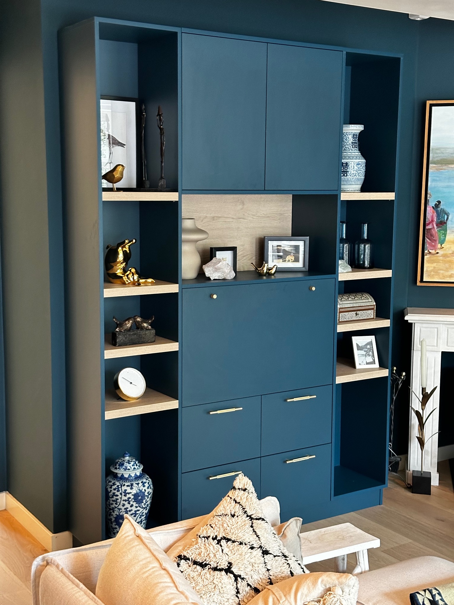 Niebieska zabudowa meblowa z półkami i szafkami, dekorowana bibelotami. Widoczny fragment sofy z poduszkami. Całość w aranżacji wnętrza.