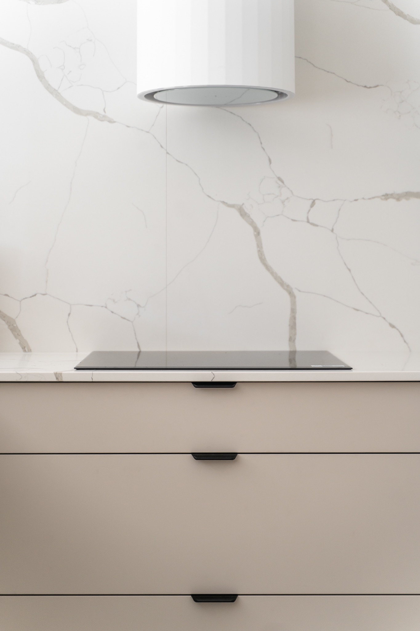 Minimalistyczna kuchnia: beżowe szafki z czarnymi uchwytami, indukcja na blacie z marmurowym wzorem i okrągły okap. Jasne, nowoczesne wnętrze.