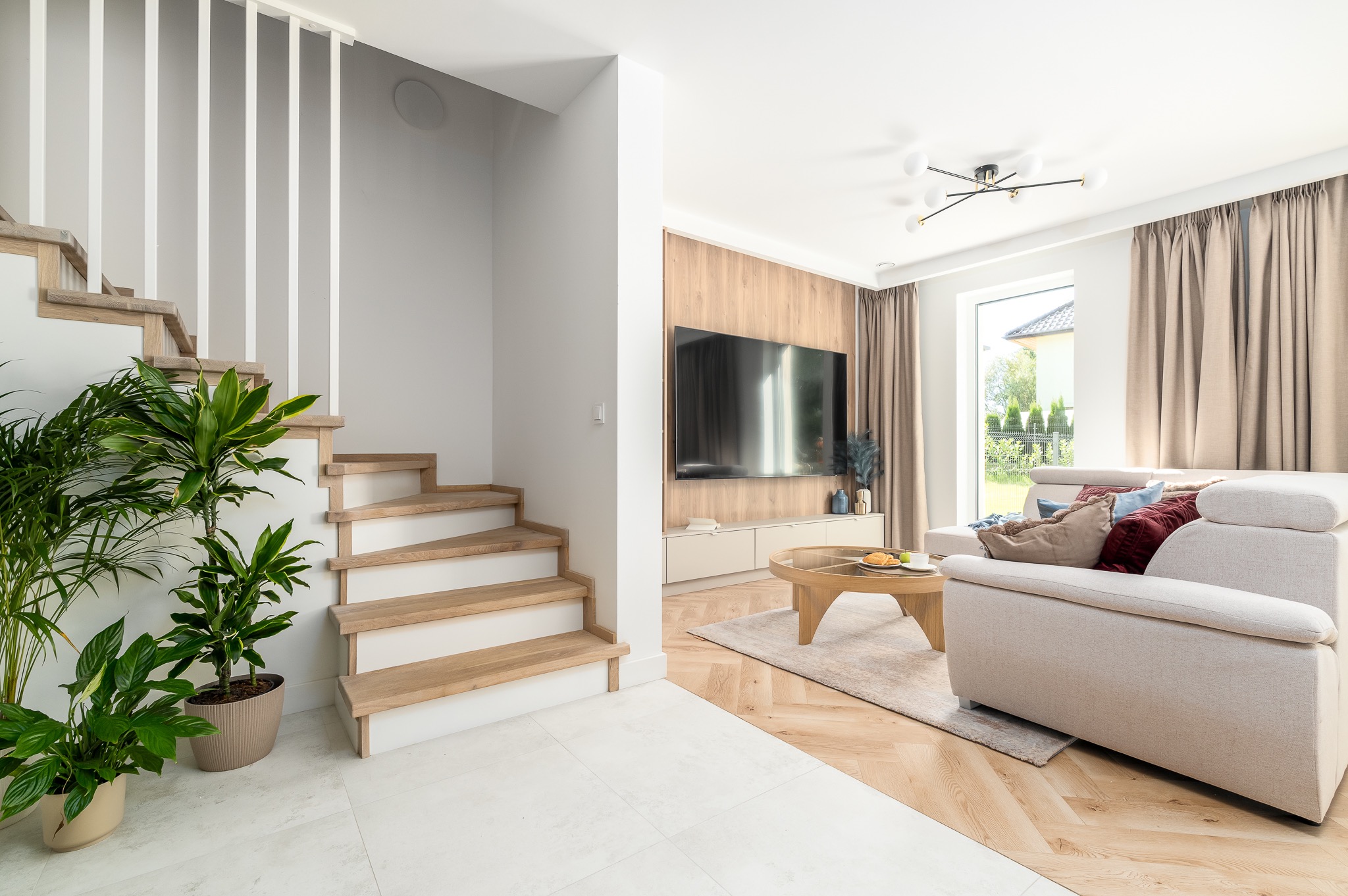Jasne wnętrze domu z drewnianymi schodami, roślinami doniczkowymi i salonem z beżową sofą. Telewizor na ścianie z drewnianą okładziną. Nowoczesny design.