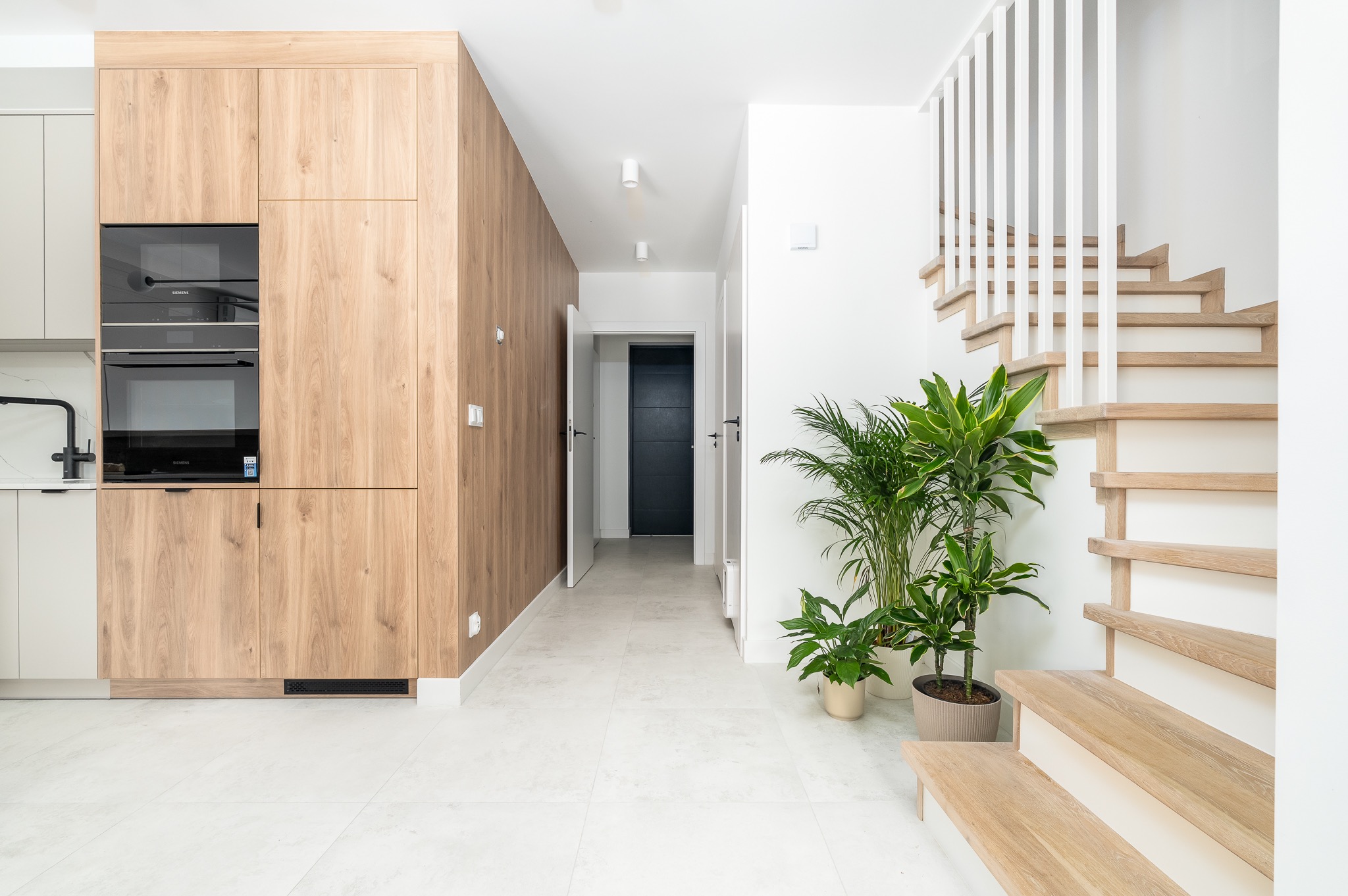 Jasne wnętrze domu z drewnianą zabudową kuchenną, schodami i roślinami doniczkowymi. Minimalistyczny design, białe ściany i podłoga. Widok na korytarz z ciemnymi drzwiami.