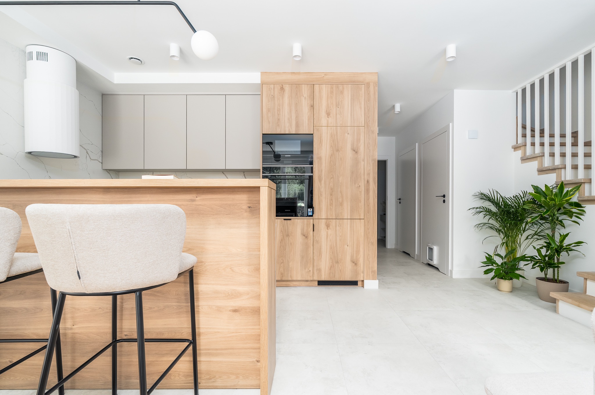 Jasna kuchnia z drewnianą zabudową i wyspą, krzesła barowe z beżową tapicerką. Minimalistyczny design, białe ściany i podłoga, roślina doniczkowa przy schodach.