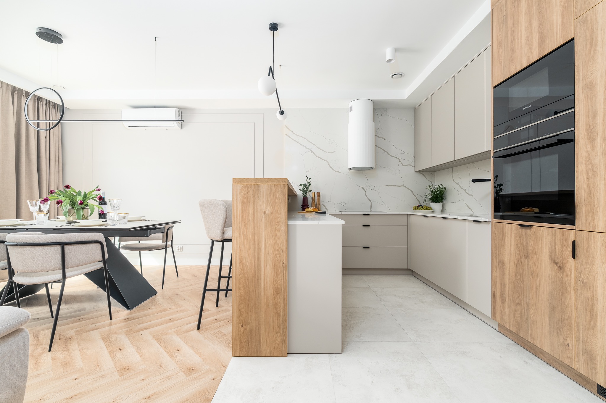 Jasna kuchnia z drewnianymi akcentami: wyspa, szafki, sprzęt AGD. Marmurowa ściana i podłoga. Stół z kwiatami w tle. Nowoczesny design, minimalistyczne oświetlenie.