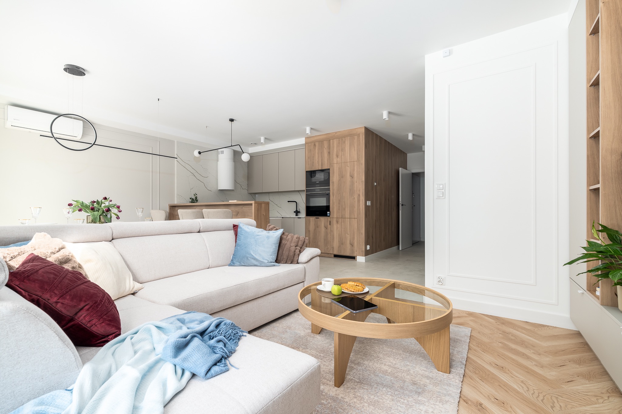 Jasne, przestronne wnętrze: salon z beżową sofą, okrągłym stolikiem kawowym i widokiem na kuchnię z drewnianą zabudową. Parkiet w jodełkę, nowoczesne oświetlenie.