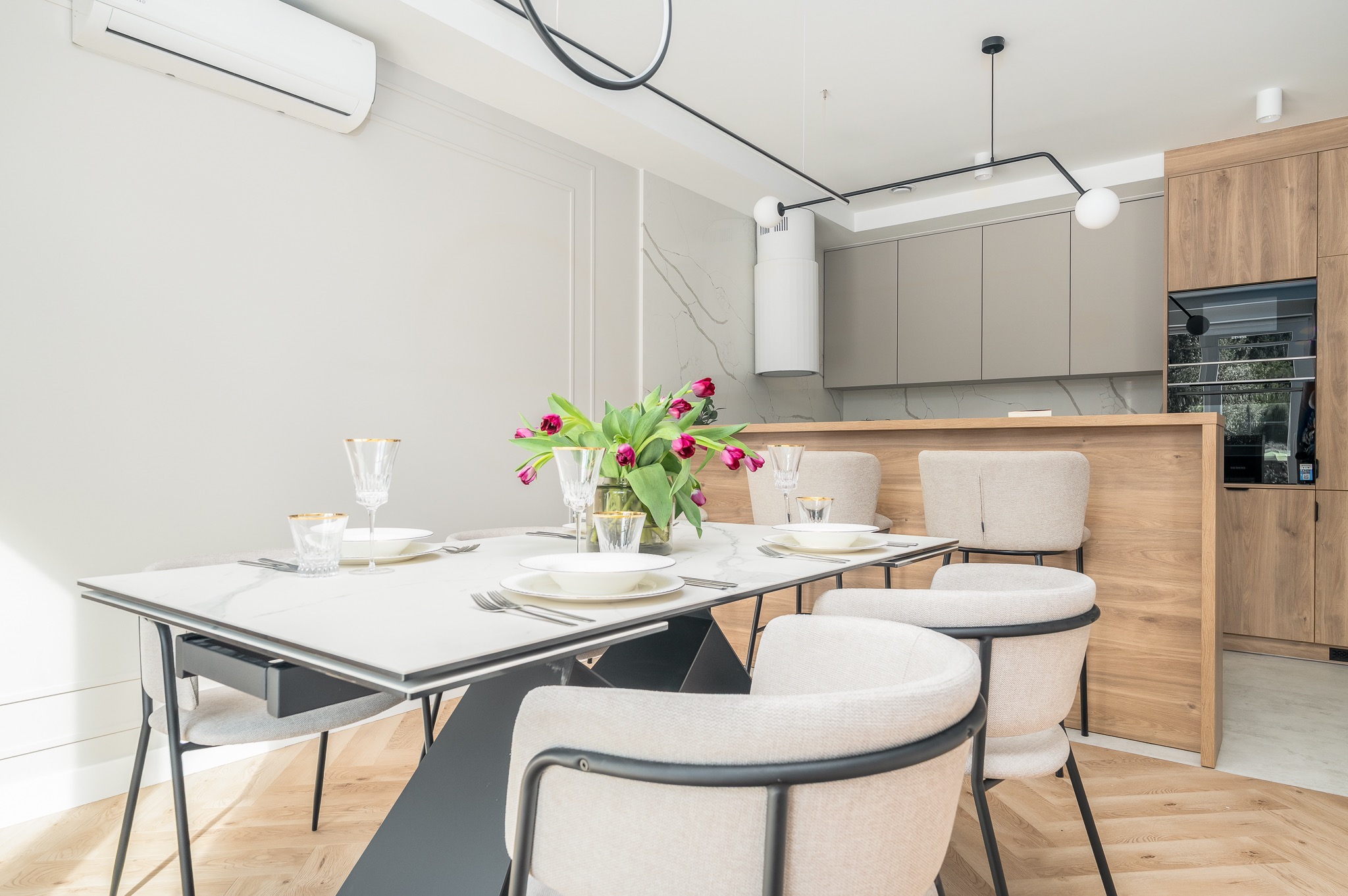 Jasna jadalnia z marmurowym stołem, kwiatami i krzesłami w stylu skandynawskim. W tle zabudowa kuchenna z drewnianymi frontami i sprzęt AGD.