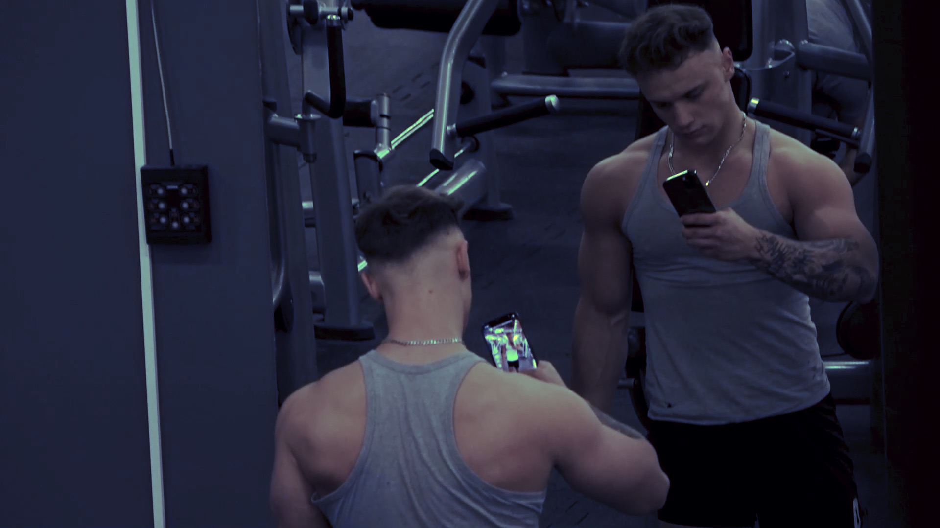 Dwóch wysportowanych mężczyzn w siłowni, jeden z tatuażem na ramieniu sprawdza coś na telefonie, drugi robi mu zdjęcie telefonem.